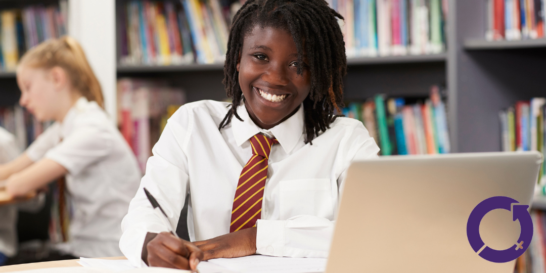 An of a teenage girl in a school uniform. She is sitting down and smiling at the camera, whilst a laptop sits in front of her. 