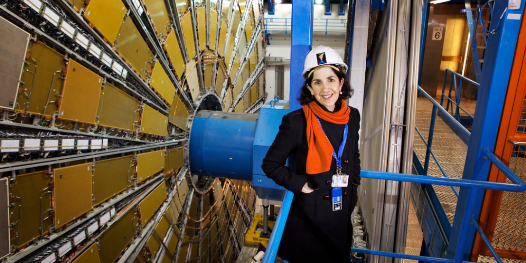 An image of famous scientist and physicist Fabiola Gianotti. She is around large, heavy machinery, smiling whilst wearing a hard hat. 